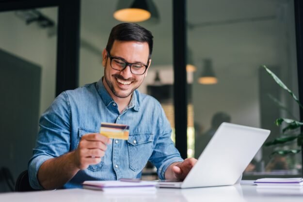 Happy cheerful smiling young adult man doing online shopping or e-shopping satisfied entrepreneur making online payment paying for service or goods self employed freelancer collecting fee